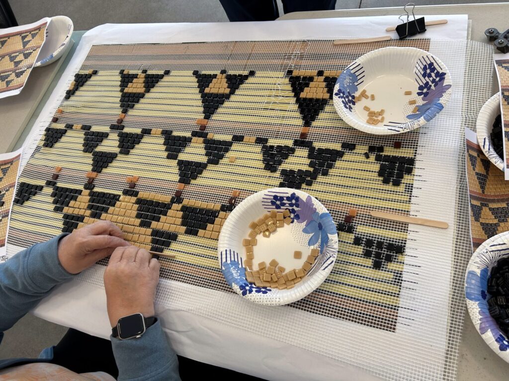 A persons hands are shown in the lower left corner of the image as they work to place tan and dark brown small square tiles into a triangular pattern on a larger canvas that will become a tile mosaic.
