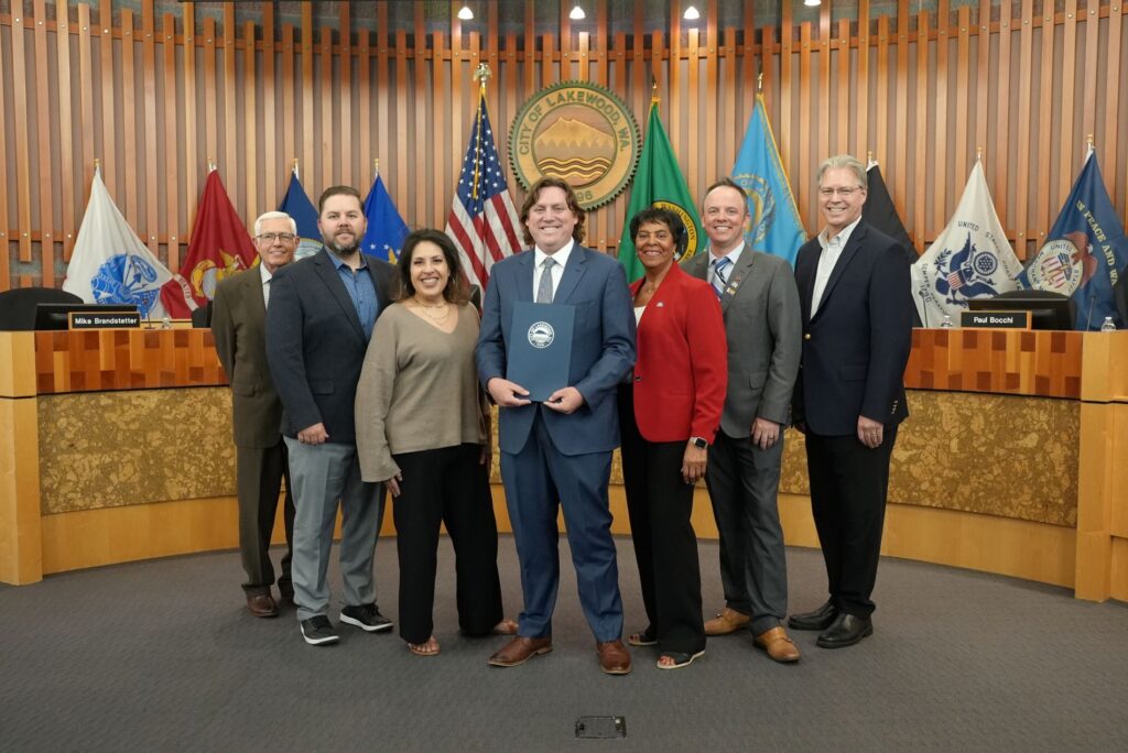 The Lakewood City Council poses with a representative from Pierce Transit who is wearing a suit and holding a proclamation. They are at the front of the city council chambers with flags in the background and the Lakewood city seal on the wall.