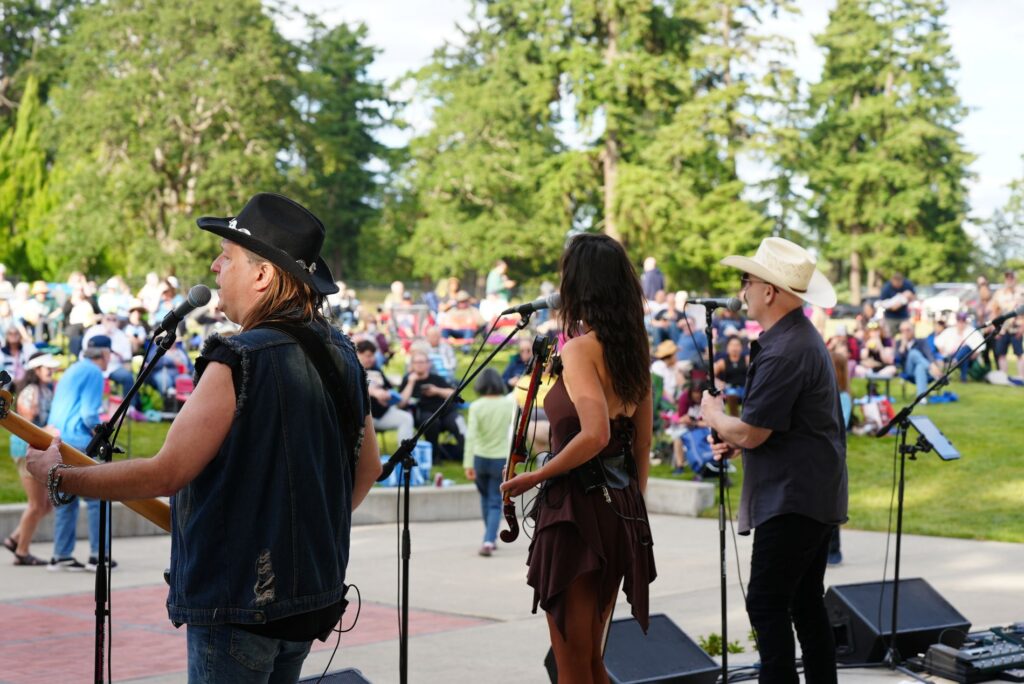 Rebel Canyon band performs on an outdoor stage in Lakewood. In the background a crowd of people is seated with tall trees behind them.