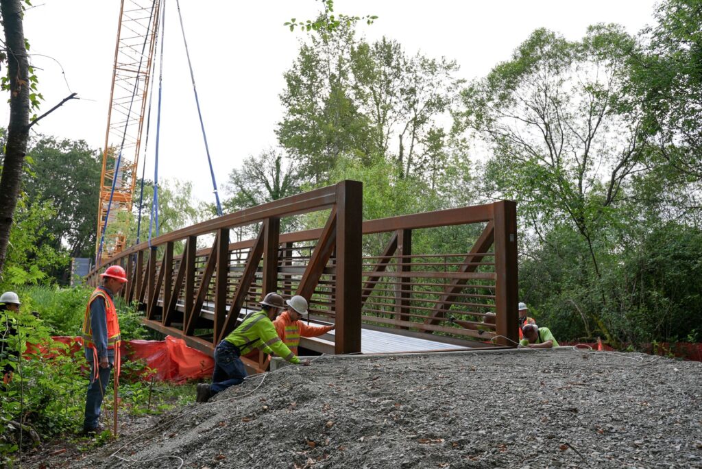 Workers in hard hats and brightly colored construction vests work around the edges of a large bridge span that is being installed with a crane at Wards Lake Park. The setting is outdoors with trees and shrubs visible.