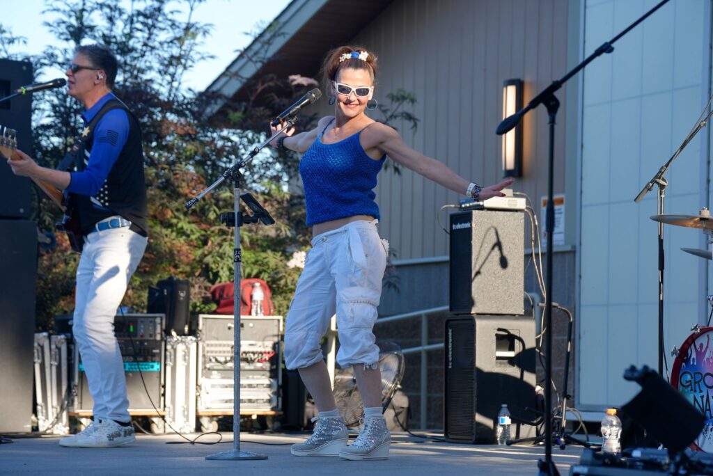 A singer performs on the Pavilion Stage for the Summer Nights at the Pavilion summer concert. She is in a blue tank top, white pants and silver sparkly shoes. She is wearing white sunglasses, has her arms out to her sides and smiles at the camera.