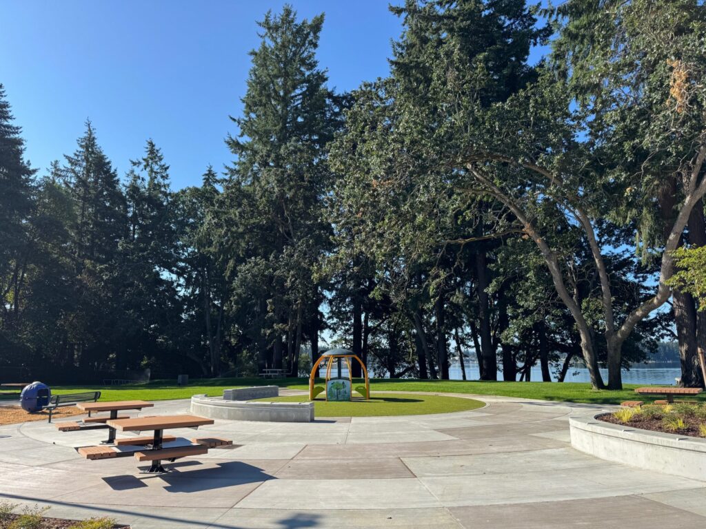 New park picnic tables are seen off to the left, a new paved gathering area is centered and tall evergreen trees are in the background of this image of American Lake Park.
