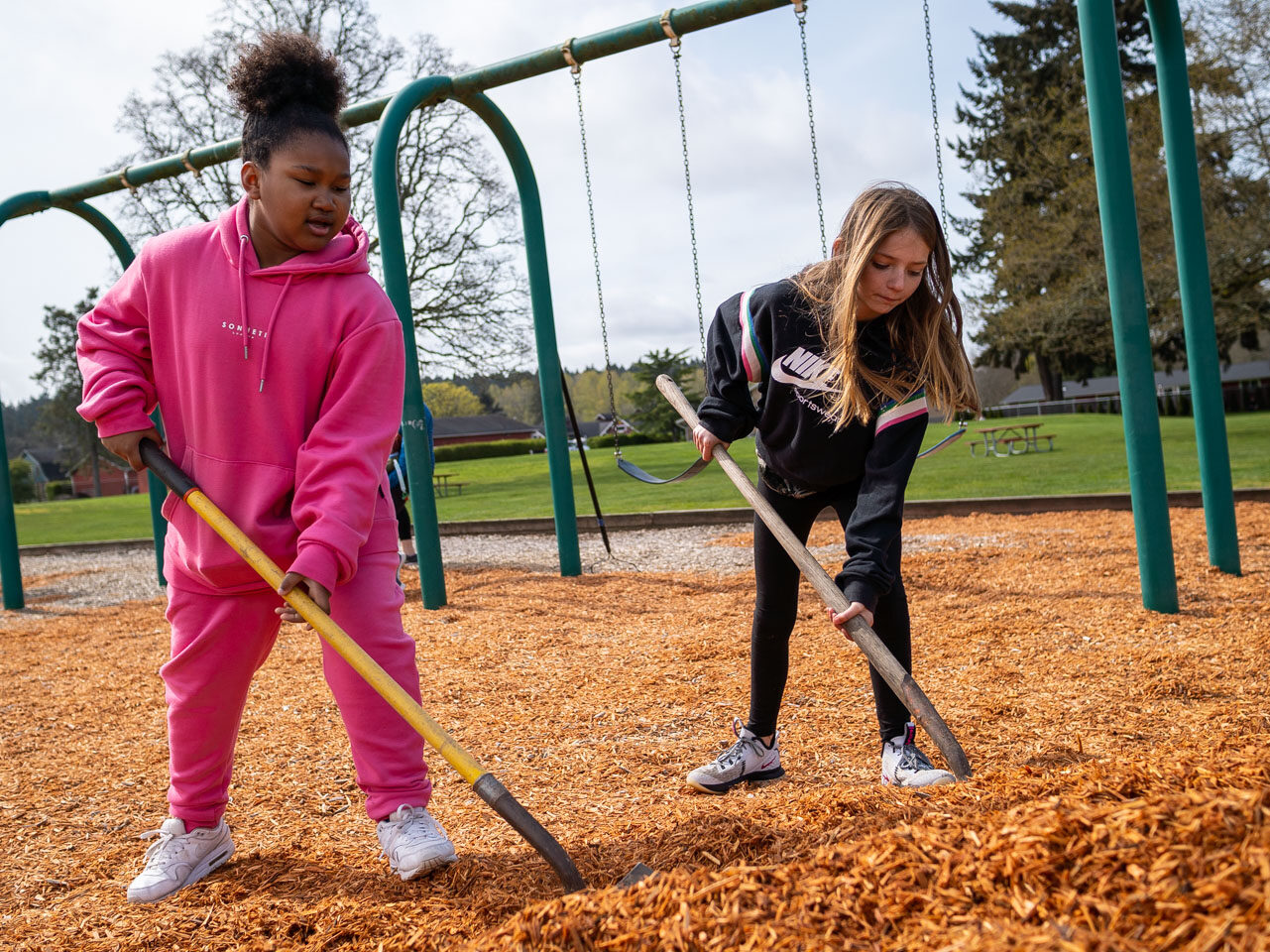 Two young people use shovels to spread woodchips at a playground during a volunteer event.