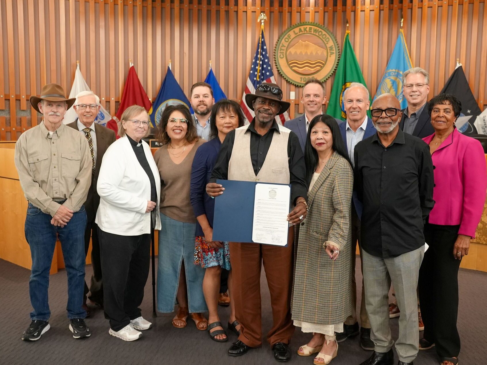 The Lakewood City Council poses with representatives from St. J-Js Episcopal Church who hold up a proclamation recognizing Juneteenth 2025. They are in Council Chambers with the city seal on the wall behind them and flags on display.