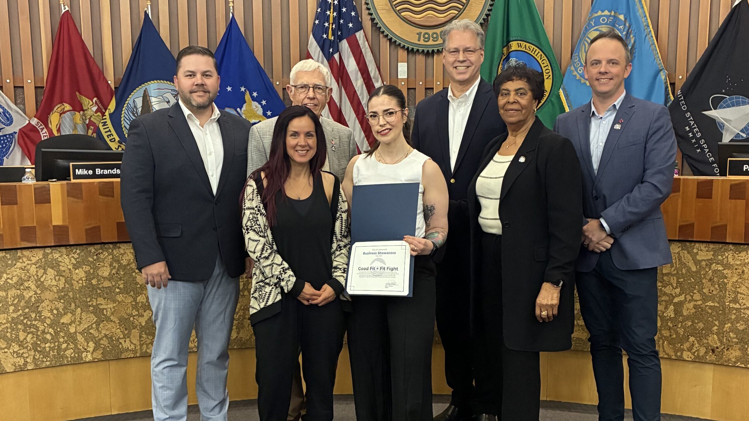 Members of the Lakewood City Council stand with business owners of Good Fit + Fit Fight, a local gym that was recognized as the May 2025 Business Showcase. They are in the Lakewood City Council chambers with flags in the background.