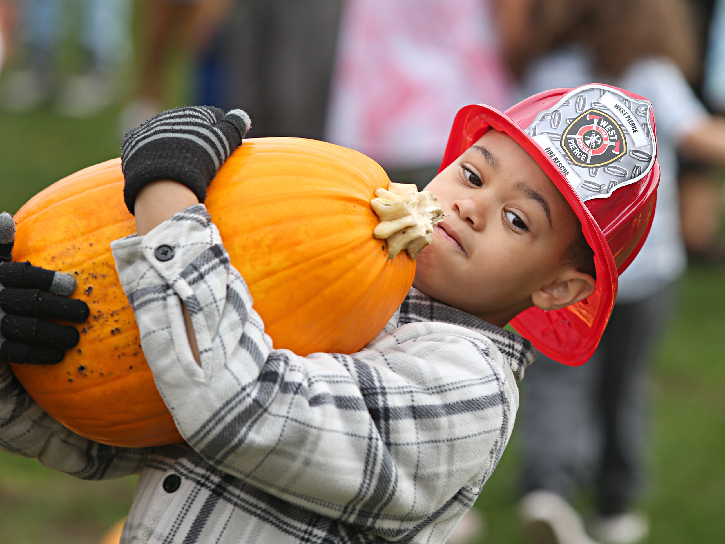 A child leans back as he holds a large pumpkin at the Lakewood Truck & Tractor Day 2023 event. He is wearing a red West Pierce Fire & Rescue plastic hat.