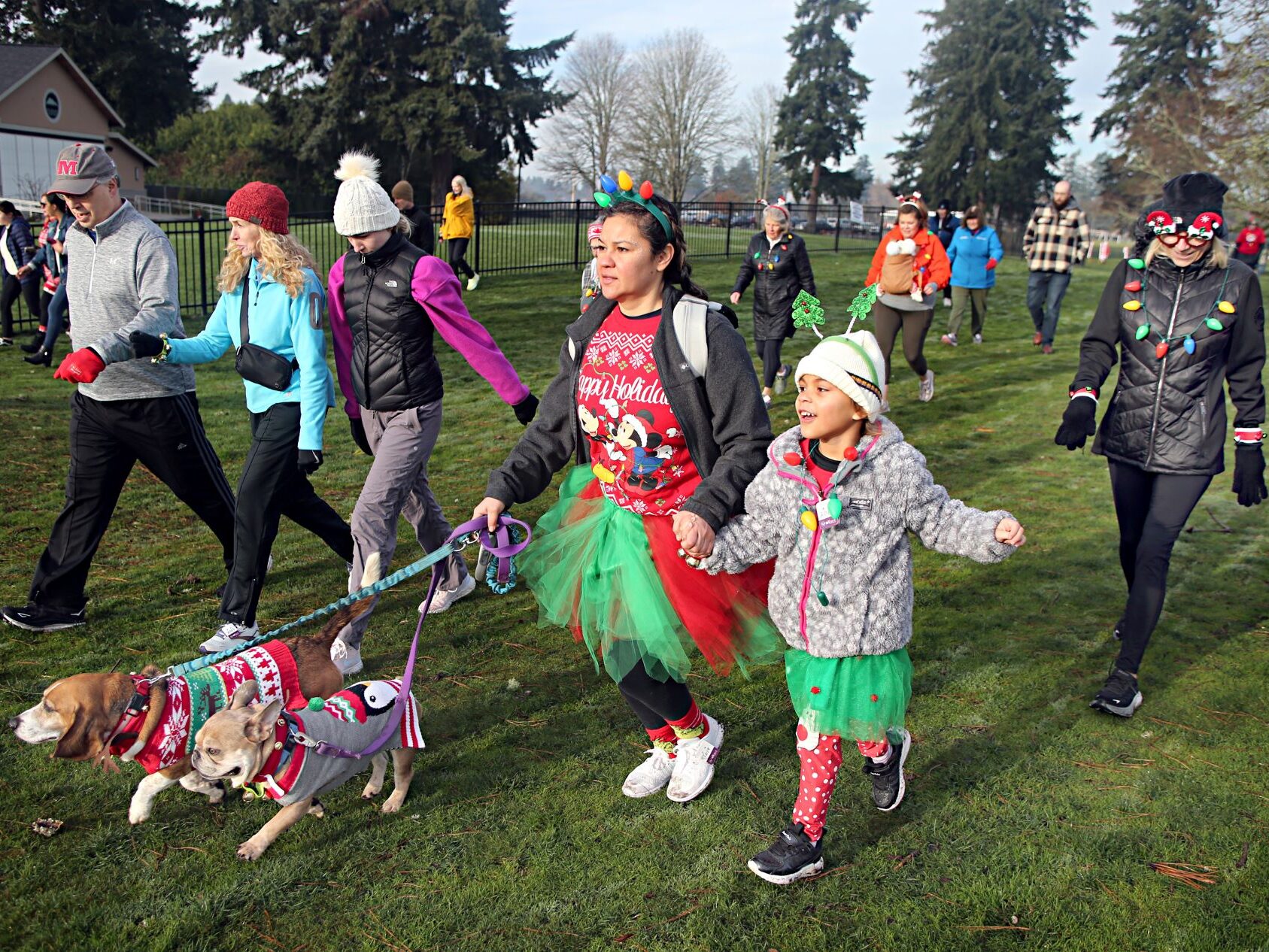People dressed in holiday costumes participate in the 2023 Jingle Bell 5k walk/run at Fort Steilacoom. A woman walks two dogs on a leash dressed in costumes.