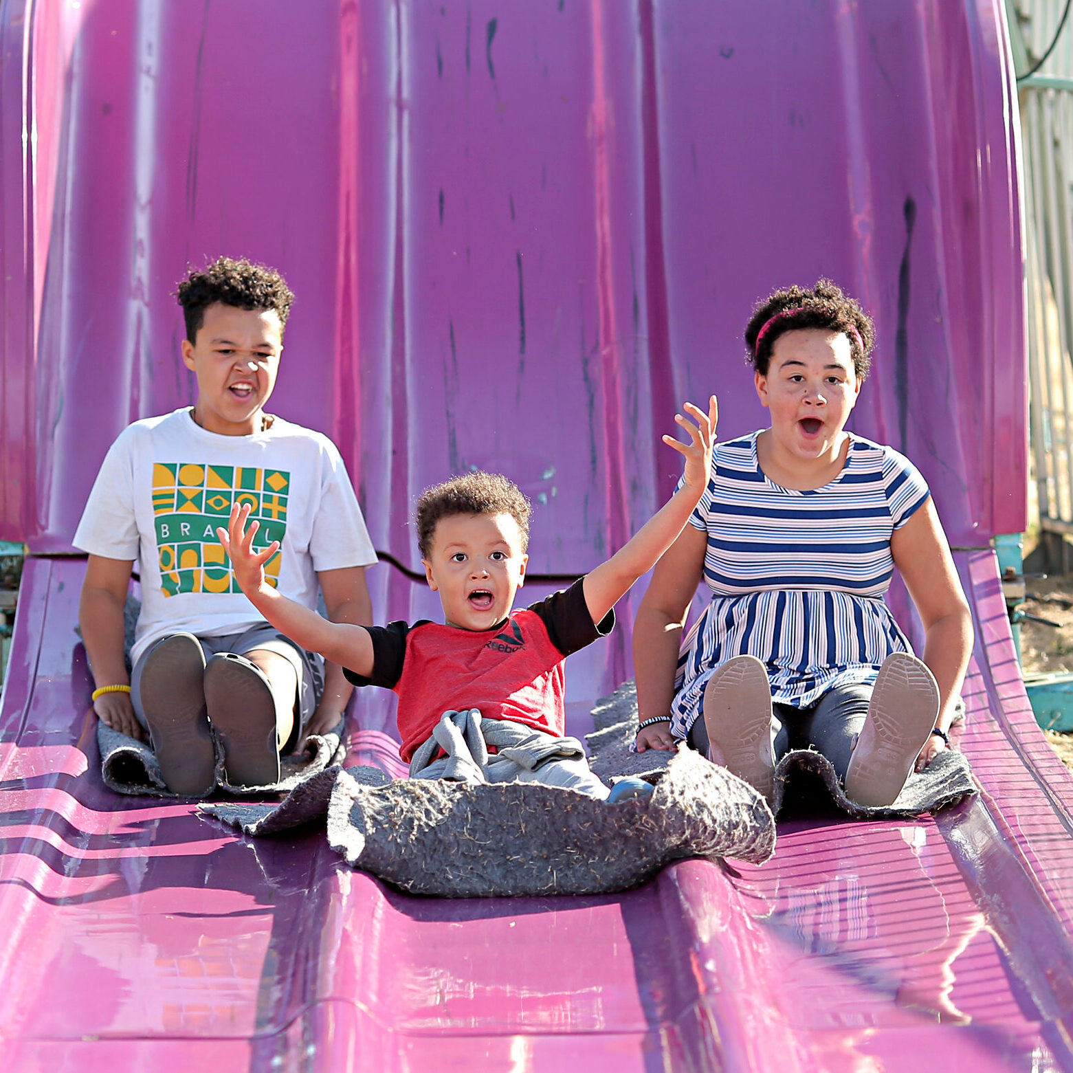 Three children come down a giant purple slide. The middle child throws their hands up in joy. The other two are smiling and excited.