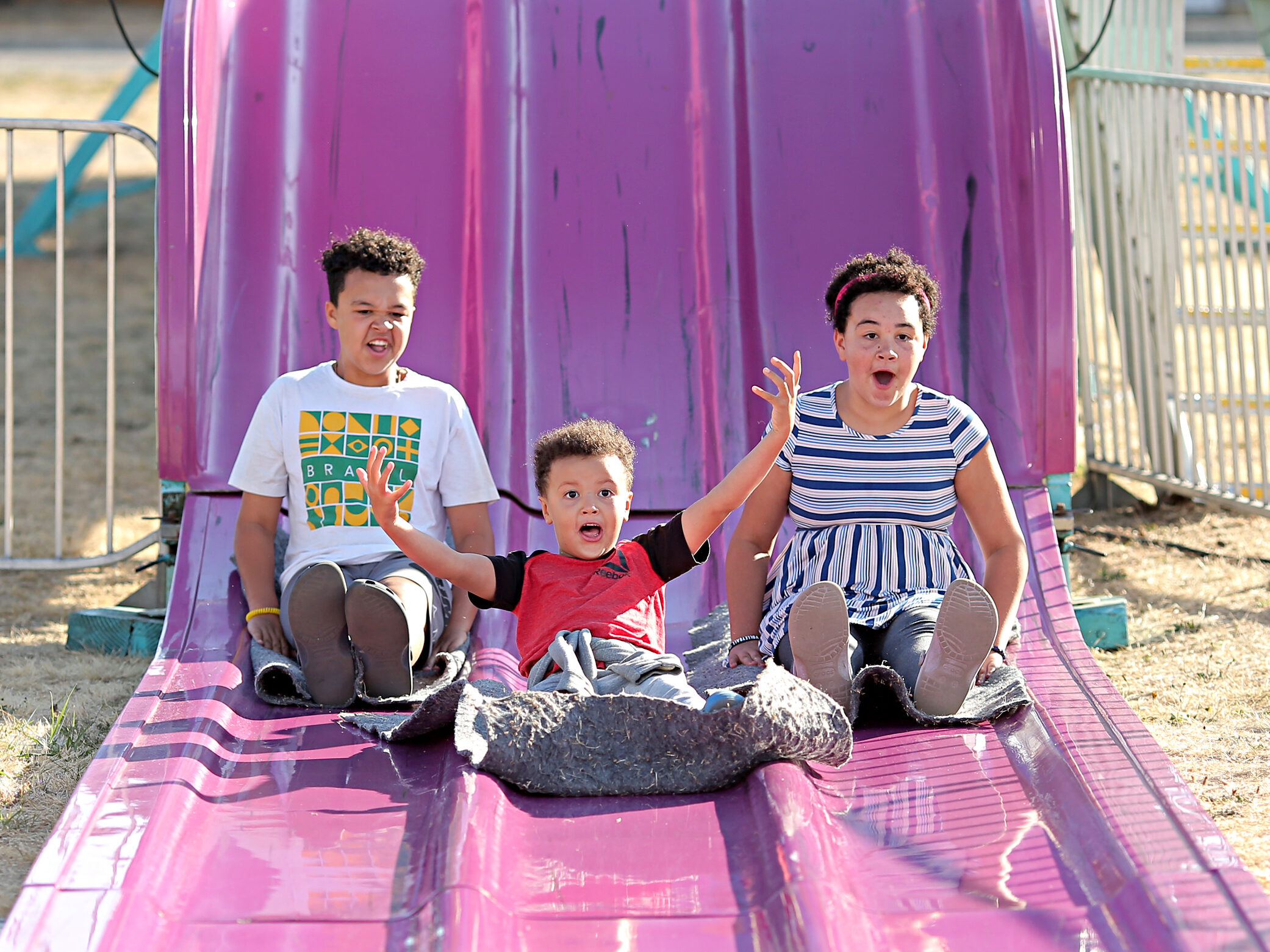 Three children come down a giant purple slide. The middle child throws their hands up in joy. The other two are smiling and excited.