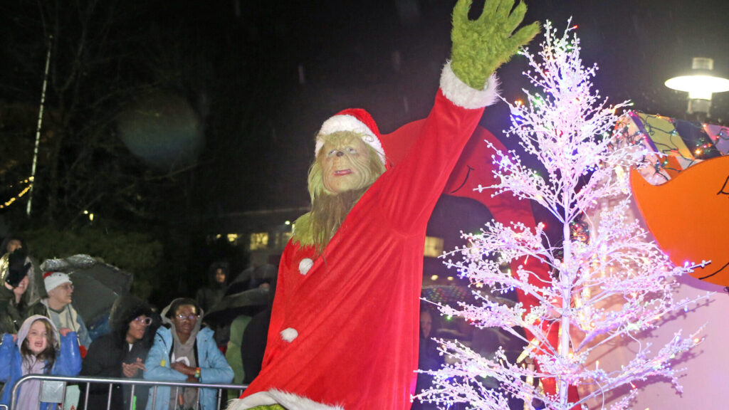 A person dressed in a Grinch costume waves to the crowd while standing on a parade float in the 2023 Holiday Parade of Lights