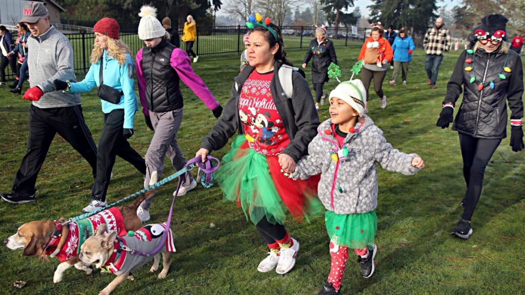 People dressed in holiday costumes participate in the 2023 Jingle Bell 5k walk/run at Fort Steilacoom. A woman walks two dogs on a leash dressed in costumes.