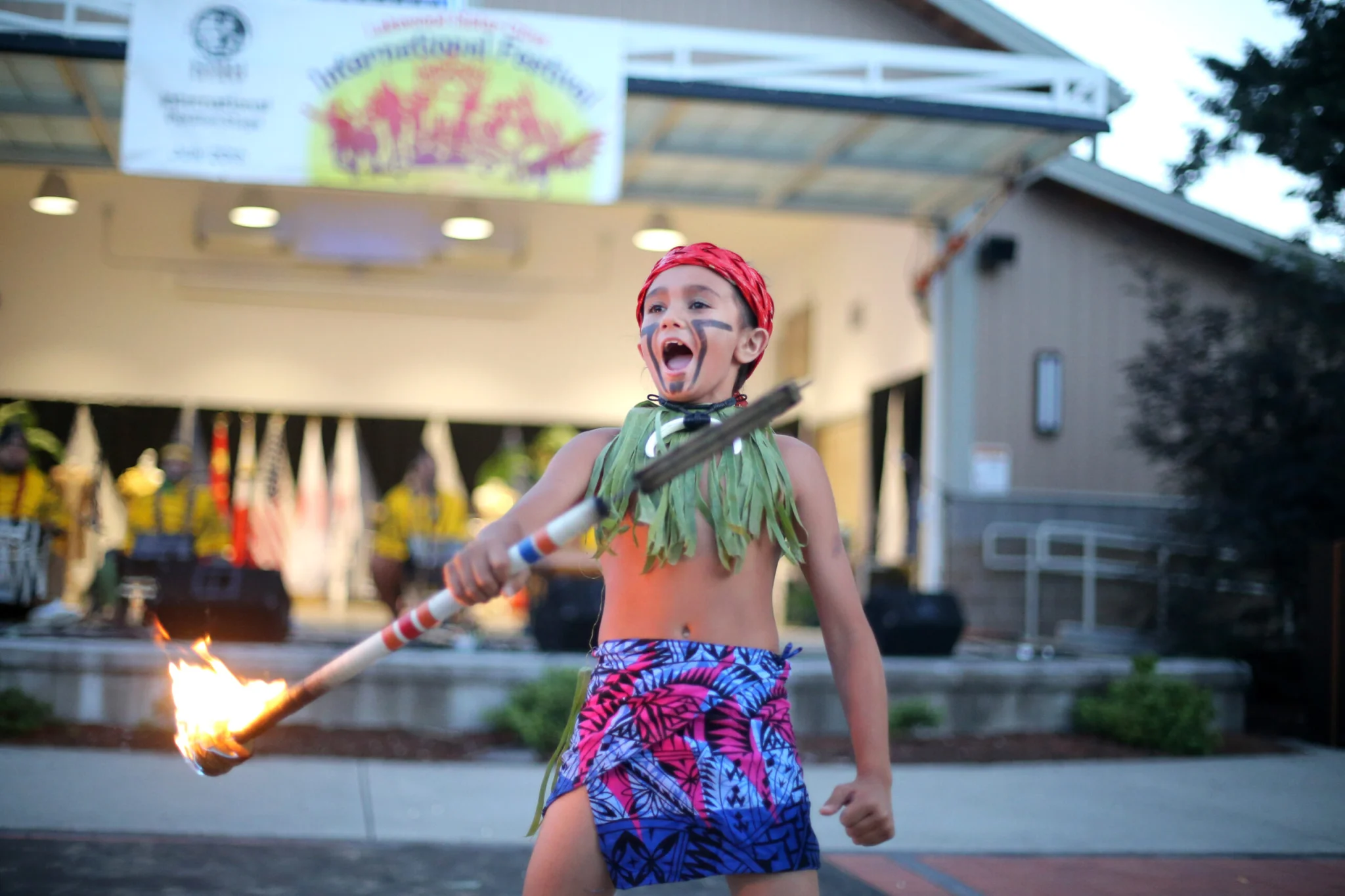 A young boy in traditional cultural gear holds a stick on fire and yells at a crowd.