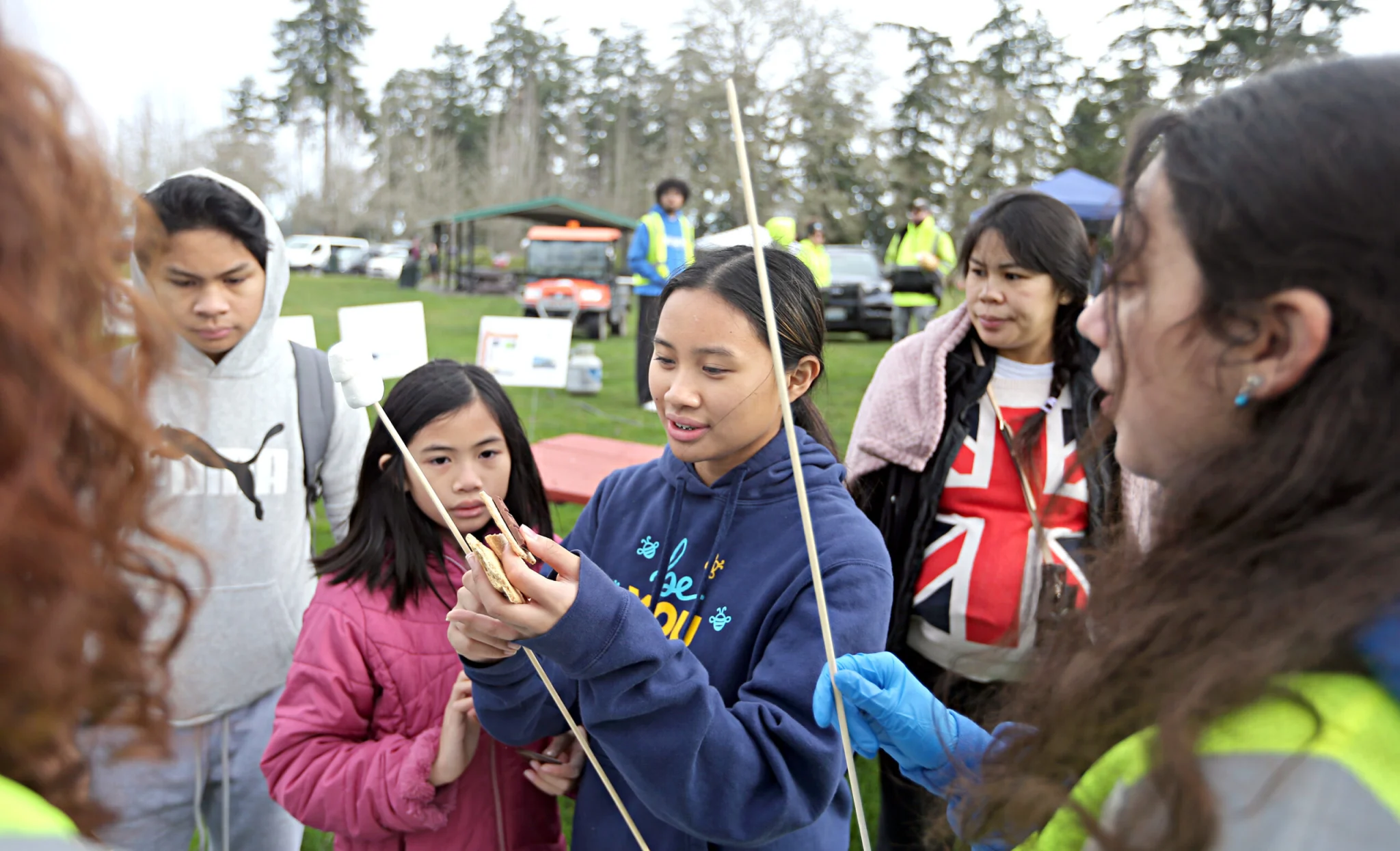 Two girls hold s'mores while a volunteer wearing a yellow vest helps them.