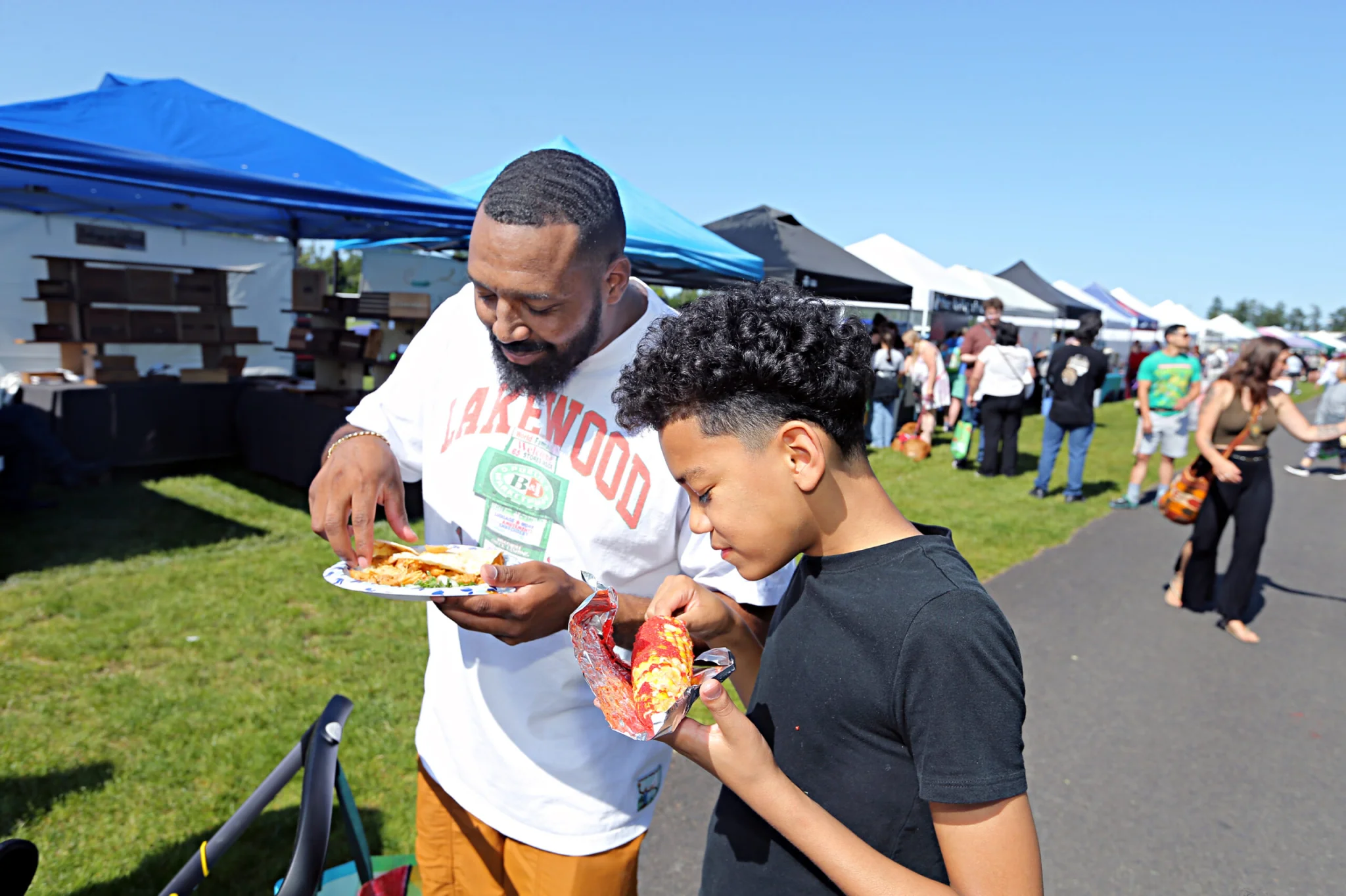 An adult and a child enjoy food from a food truck at the Lakewood Farmers Market. They are standing on the main paved path in the market with booths in the background